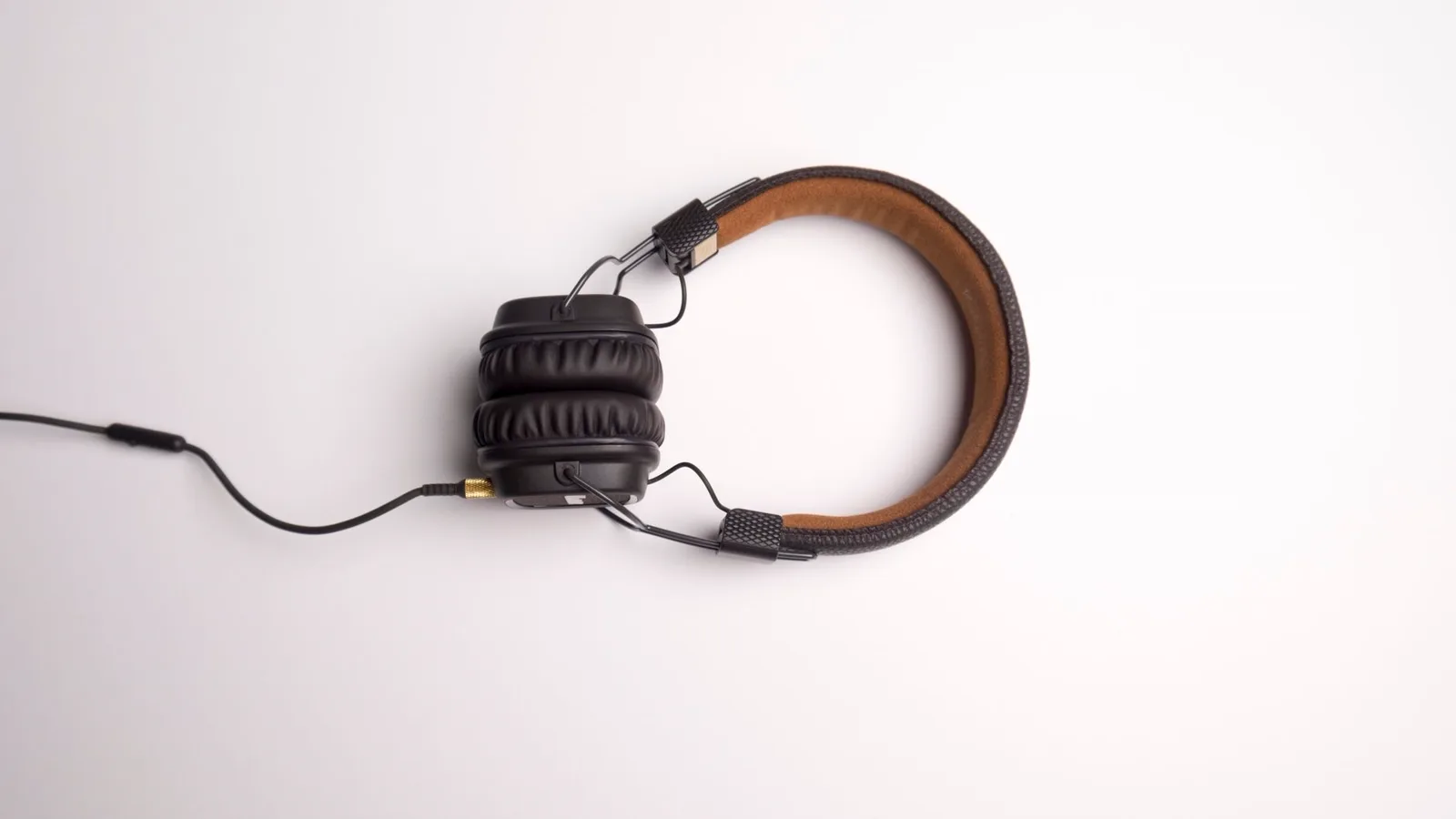 Fone de ouvido com fio circumaural em cores neutras sobre escrivaninha de escritório com monitor, teclado e café ao fundo; luz natural de janela; foco no cabo e conector do fone.